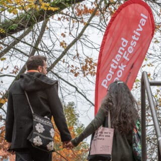 Two individuals, holding hands and carrying book bags, walking in front of a Portland Book Festival sign